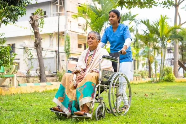 Wide shot of Nurse taking senior woman on walk while on wheelchair at hospital garden - concept of caretaker, disability and healthcare — Stock Photo, Image