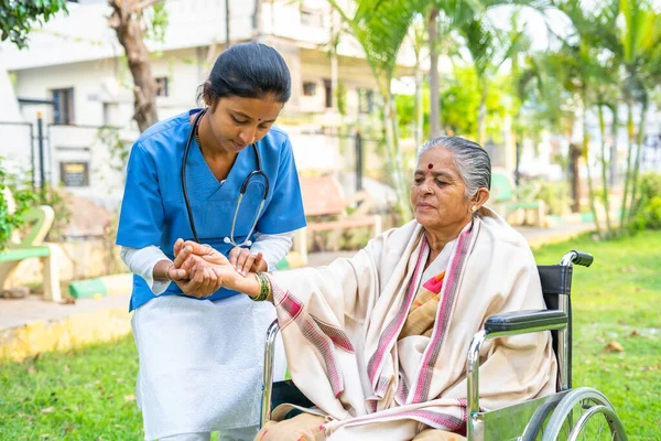 Doctor checking pulse rate of disabled woman while on wheelchair at park - concept of medical care, caretaker and professional occupation — Stock Photo, Image