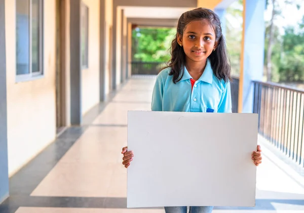 Χαμογελώντας κορίτσι παιδί δείχνει λευκό άδειο πίνακα κοιτάζοντας κάμερα στο διάδρομο του σχολείου - έννοια της εκπαίδευσης, διαφήμιση και προώθηση — Φωτογραφία Αρχείου
