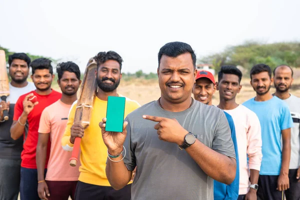 Игроки в крикет показывают мобильный телефон с зеленым экраном, указывая пальцем на камеру на детской площадке - концепция продвижения приложений, рекламы и дружбы — стоковое фото