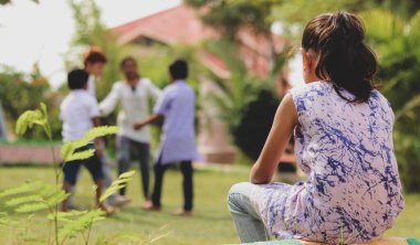 Üzgün Genç Küçük 'ün parkta tek başına oturup parkta oynayan diğer çocuklara bakması - yalnızlık, depresyon, reddedilme ve kabul edilmeme kavramı.