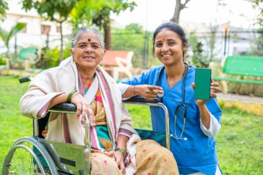 Parkta tekerlekli sandalyede oturan hasta yeşil ekran cep telefonu gösteriyor. Teknoloji kavramı, sağlık uygulaması tanıtımı ve reklam..