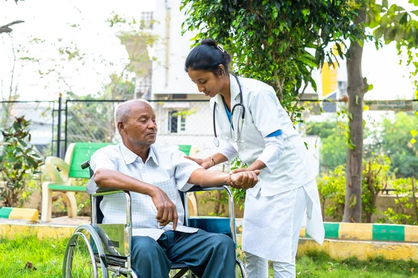 Doctor helping senior old patient to stretch hands during morning exercise while on wheelchair at hospital park - concept of physio, caretaker and therapy or treatment. — Stock Photo, Image