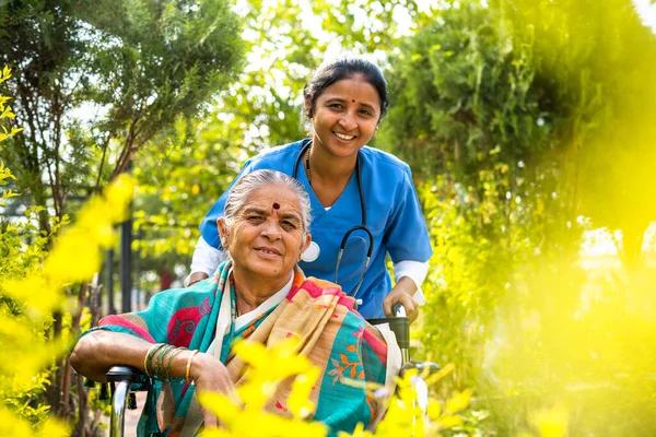 Happy smiling Nurse taking senior woman on walk while on wheelchair at hospital garden by looking at camera - concept of caretaker, disability and healthcare — Stock Fotó