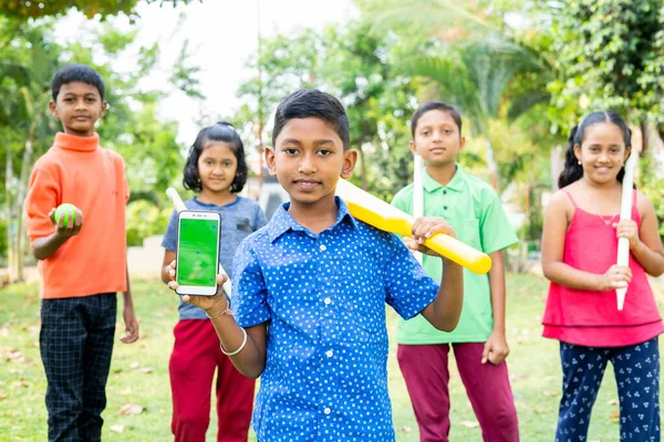 Smiling kid showing green screen mobile phone with cricket bat by looking at camera with friends at park - concept of training, advertisement and promotion — Stock Photo, Image