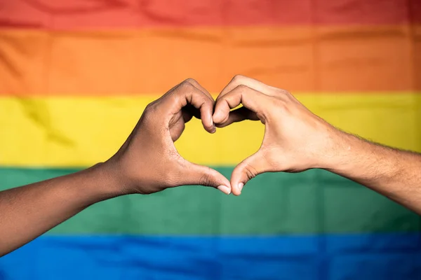 Zbliżenie ujęcie rąk uprawiających miłość lub symbol serca przed dumą lub tęczową flagą - koncepcja gejowskiej pary miłość, solidarność i święto miesiąca dumy. — Zdjęcie stockowe
