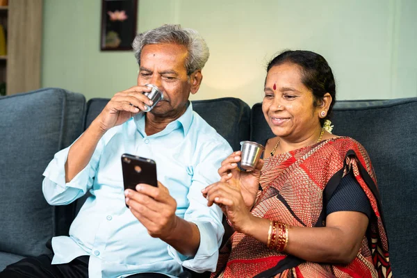 上了年纪的夫妇一边在沙发上喝茶一边用手机笑 亲情和幸福的概念 — 图库照片