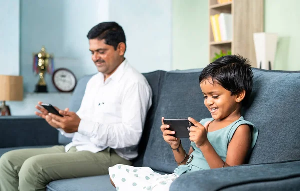 Father and daughter on sofa separately busy using mobile phone at home - concept of digital divide, social media and technology addiction.