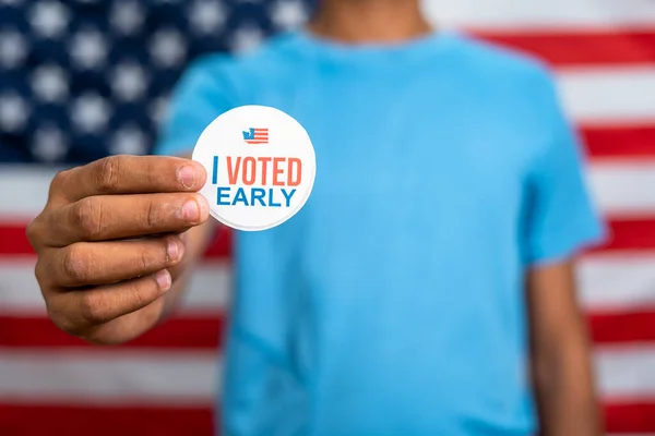 close up shot of young man showing I voted early stick in front of US american flag - concept of early voting or polling