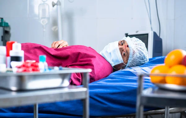 Senior sick patient on hospital bed with medical face mask and operation gown looking at camera - concept medical treatment, disease and recovery.