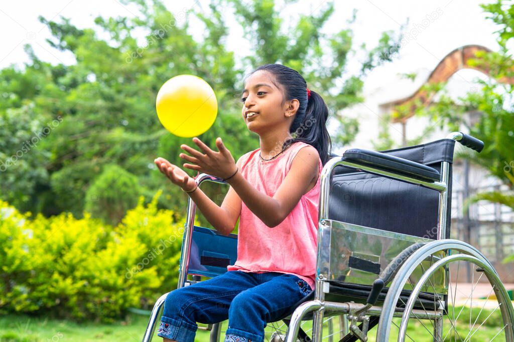 concept of happiness, freedom and enjoyment showing by Smiling alone girl kid with disability playing with ball while on wheelchair at park