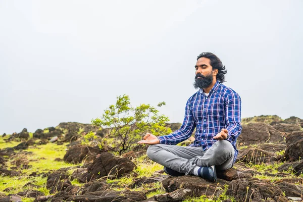 Young beard man with eyes closed doing meditation or yoga on top of hill - concept of self caring, health care and wellness.