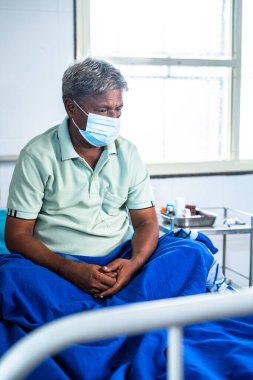 Vertical shot of thoughful elderly man with medical face mask sitting on hospital bed - concept of worried, loneliness and depression.