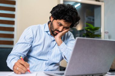 Ofis masasında not alarak dizüstü bilgisayar başında çalışan yorgun genç adam - iş sıkıntısı, rutin iş hayatı ve yorgunluk kavramı