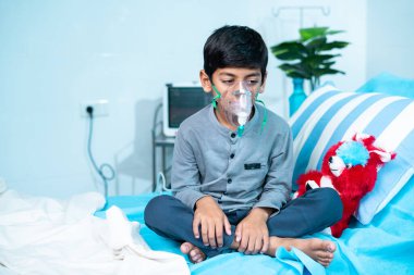 Worried sick kid with oxygen mask sitting on bed at hospital ward - concept of breathing problems, respiratory infection and health care treatment