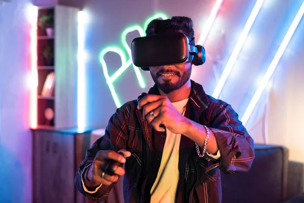 Young man on neon background playing video game virtual reality or VR headset at home using gamepads - concepts of metaverse, cyberspace and futuristic.