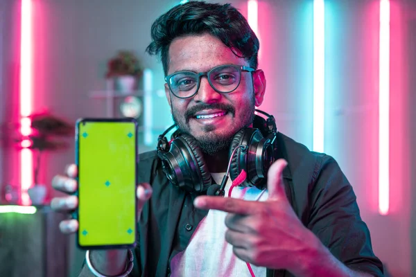 Professional gamer with headset showing green screen mobile phone by pointing finger while looking camera at home - concept of champion,app advertisement and champion