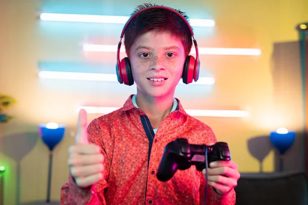 young Teenager kid with headphones holding gamepad by showing thumbs up by looking at camera at home - concept of live streamer and gaming vlogger