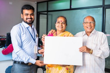 Happy Smiling banker with senior couple showing white empty board by looking camera at bank - concept of advertisement, banking promotion and customer service