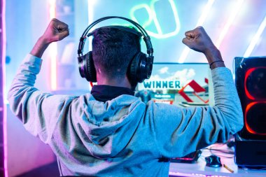 shoulder shot of gamer with headphones celebrating win by raising hands while playing live video game on computer at home - concepts of champion, successful and gaming tournament