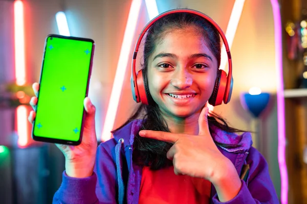 Happy teenager girl kid with headset showing green screen mobile phone by pointing finger by looking camera at home - concept of app advertisement, entertainment and promotion