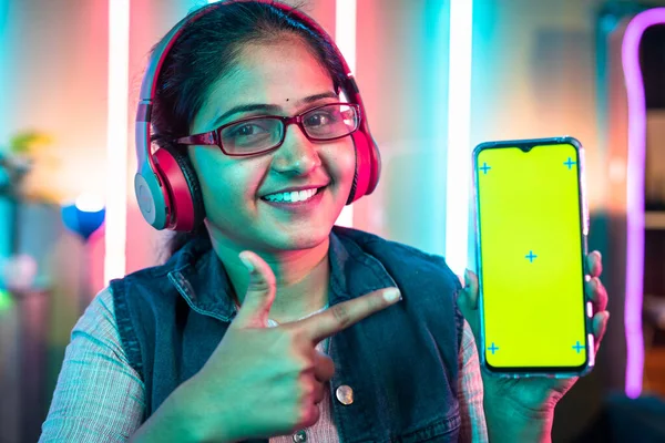 Smiling woman with wireless headset showing green screen mobile phone by pointing finger while looking camera at home - concept of gaming advertisement, app promotion and technology.