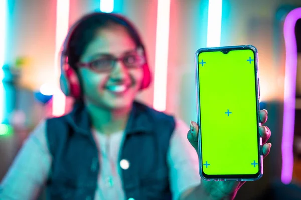 focus on mobile phone, Smiling girl showing green screen mobile phone by looking camera on neon gaming background - concept of gaming advertisement, app promotion and technology