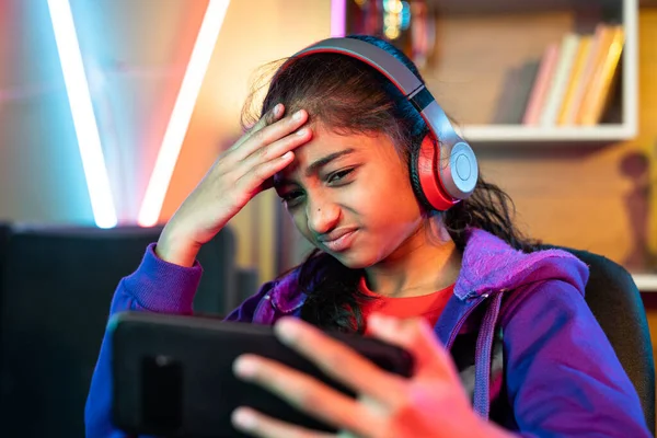 Sad girl with headphones losing game while playing video game on mobile phone at home - concept of competition, technology and entertainment.