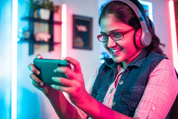 happy smiling girl with headphones and eyeglass playing online video game on mobile phone at home - concept of live streamer, holydays and cyberspace.