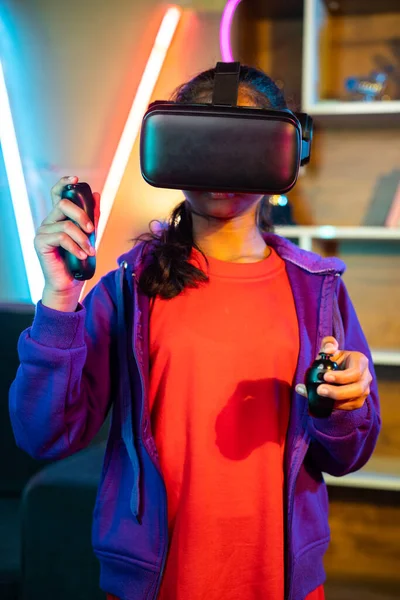 vertical shot of Young teenager girl playing video game on virtural reality or VR headset using joystick at home on neon background - concept of metaverse, cyberspace and futuristic lifestyle.