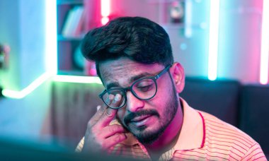 Evdeki bilgisayarda oyun oynarken göz tahrişi, oyun bağımlılığı, yorgunluk ve rekabet konsepti nedeniyle gözlerini ovuşturan genç adamın yakın çekimi.