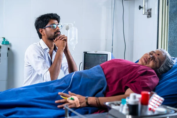 Worried indian son praying god for sick mother for speed health recovery at hospital - concept of healthcare, unconditional bonding and treatment