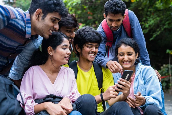 group of indian young student using social media or watching entertainment on mobilephone during free time at collage campus - concept of leisure activity, togetherness and eduaction