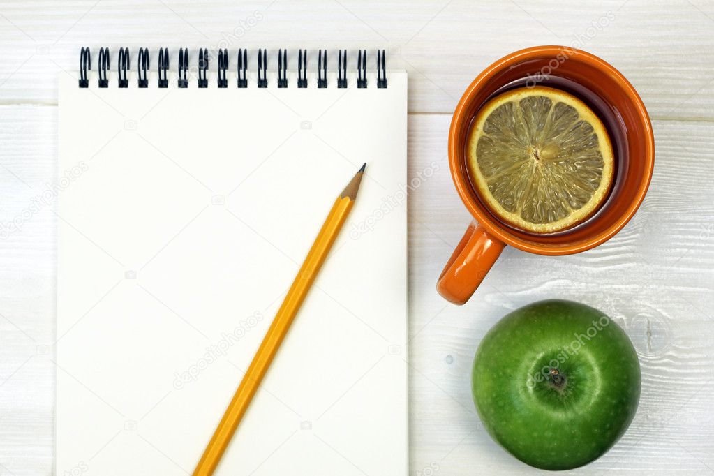 Tea, green apple, pencil and notebook on a white wooden surface
