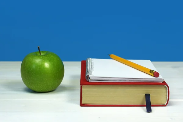 Green apple and book, with notebook and pencil on white wooden surface