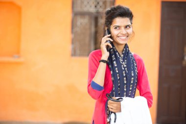 Genç gülümseyen Hintli kadın doktorun portresi akıllı telefon ya da cep telefonuyla konuşuyor, steteskop ve önlükle turuncu kırsal arka planda pozitif duygular var. Sağlık ve Tıp Konsepti.