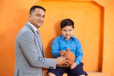 Küçük çocuklu Hintli adamın portresi kilden para kutusuna para koyuyor erkek banka müdürü ve çocuk elinde geleneksel domuz kumbarası ya da Gullak, para, yatırım ve bankacılık kavramından tasarruf etmeyi öğretiyor.