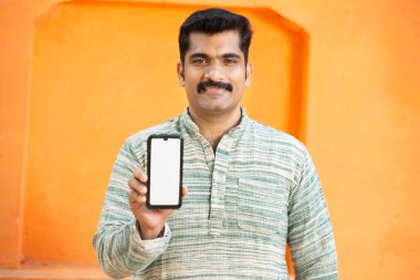 Geleneksel Hintli genç adam boş telefon görüntüsü, Bıyıklı Erkek reklam ve markalaşma için boş akıllı telefonu turuncu arka planda gösteriyor, online ödeme uygulaması, seçici odaklanma.