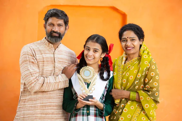 Young happy indian daughter with her father and mother holding winning prize ,Cheerful female standing with parents showing trophy against orange background. support girl child.celebrating victory.
