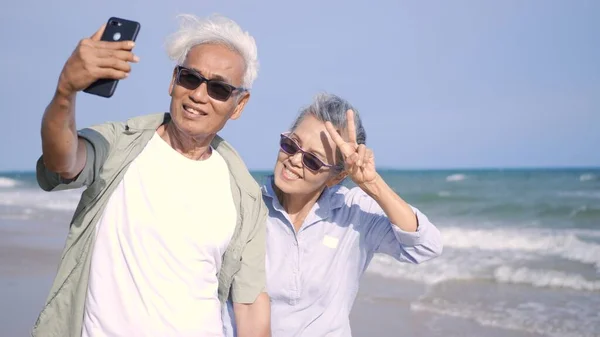 아시아인 남녀들은 웃으며 해변에서 스마트폰으로 사진을 낭만적 노인들은 휴가를 즐기고 — 스톡 사진