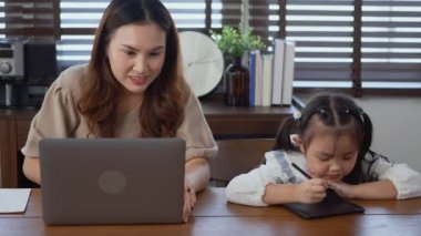 Asyalı genç anne evde dizüstü bilgisayarla çalışıyor ve kızının ev ödevlerini yapmasına yardım ediyor, bekar anne evden çalışıyor ve kızına masa başında ödev yaparken yardım ediyor.