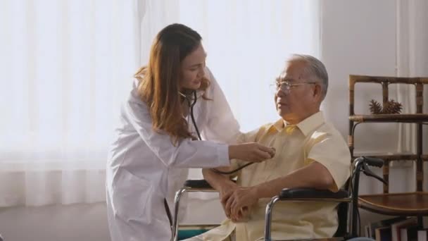 女性看護師の医師は 車椅子で座っている間に高齢者や高齢者の男性を検査する白い制服心臓科医を着用自宅で聴診器を使用してハートビートをチェック 健康訪問者の概念 — ストック動画