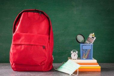 Ön okul sırt çantası ve maske takan aksesuarlar yeşil karatahtanın masasında, okul çantasındaki öğrenci çantası, okula dönüş eğitimi yeni normal salgın COVID-19 veya koronavirüs.