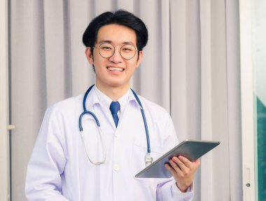 Mutlu Asyalı genç doktorun portresi. Üniformalı ve steteskoplu yakışıklı adam. Elinde akıllı dijital tablet var ve kameraya bakıyor.