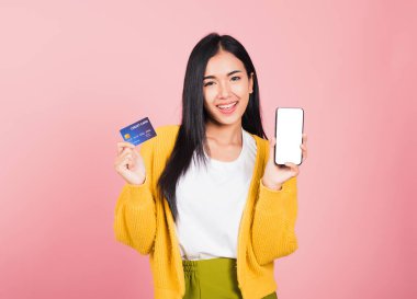 Mutlu bir Asyalı portresi güzel tatlı bir genç kadın gülümseyen cep telefonu ve plastik banka kartı, stüdyo resmi pembe arka planda, akıllı telefon kullanan bir kadın online alışveriş yapıyor.