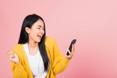 Mutlu bir Asyalı portresi güzel, güzel bir genç kadın elinde cep telefonuyla gülüyor, stüdyo çekimleri pembe arka planda, komik akıllı telefon kullanan bir kadın kazanan hareketi yapıyor.