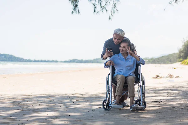 快乐的亚洲老年妇女坐在轮椅上 丈夫是一个坐在轮椅上的智能手机使用者 在海滩上自费 退休夫妇的概念 — 图库照片