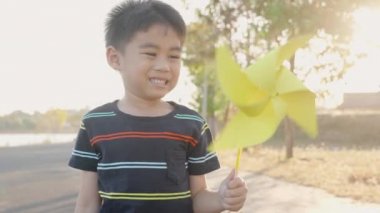 Asyalı çocuk fırıldak ile oynuyor, buğday tarlasında gülümseyen küçük çocuk küçük rüzgar tekerleği ya da yaz günü parkta elinde yel değirmeni oyuncağı tutuyor.