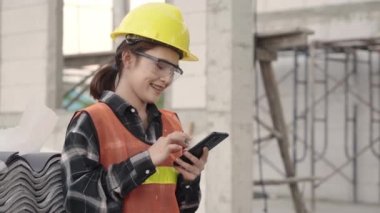 Ev inşaatında çalışırken planlama ve geliştirme projelerine takımla birlikte akıllı telefon uygulaması kullanarak koruyucu miğfer ve üniformalı Asyalı mimar mühendis kadın.