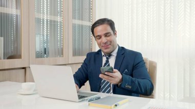 İşadamı ofisteki masa başı başarı kutlamalarında dizüstü bilgisayarını kullanarak oturuyor, gündelik ofiste mutlu bir iş adamı gibi gülümsüyor ve dizüstü bilgisayarla çalışırken telefonla konuşuyor.